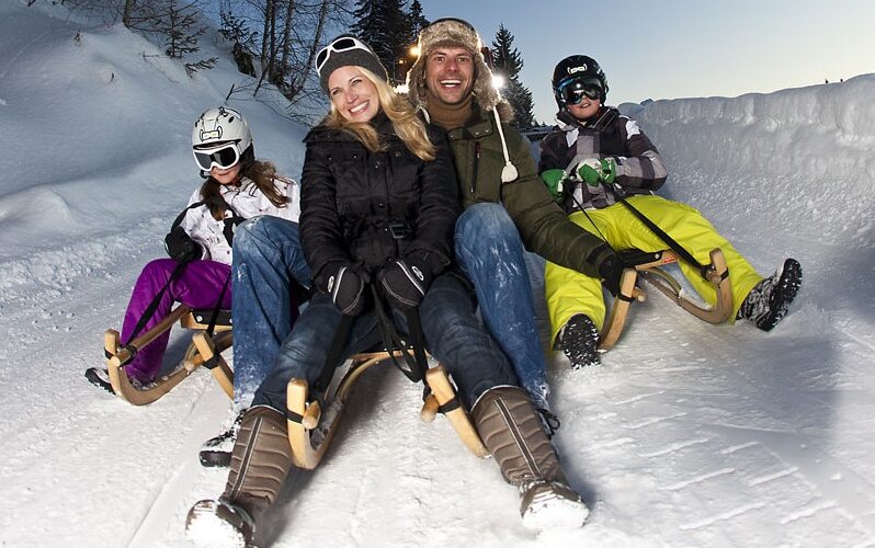 Eine vierköpfige Familie in Winterkleidung und mit Helmen fährt in der Abenddämmerung auf Holzschlitten einen verschneiten Hügel hinunter.