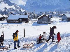 Eine Gruppe von Menschen geht durch ein verschneites Dorf, einige ziehen Kinder auf Schlitten, mit Holzhäusern und Bergen im Hintergrund.