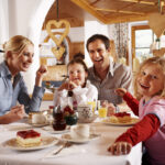 Eine vierköpfige Familie sitzt an einem Tisch in einem gemütlichen Restaurant, lächelt und genießt gemeinsam Kuchen und Getränke. Die Umgebung ist mit Holzdekor und herzförmigen Ornamenten geschmückt.