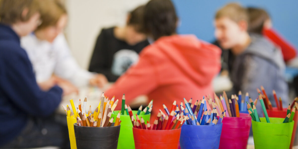 Im Vordergrund sind bunte Becher mit Bleistiften und Kugelschreibern zu sehen, während im unscharfen Hintergrund eine Gruppe von Menschen zusammensitzt.