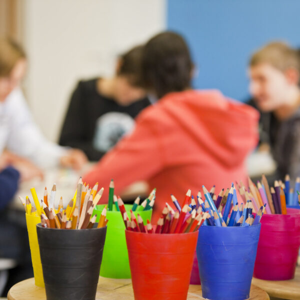 Im Vordergrund sind bunte Becher mit Bleistiften und Kugelschreibern zu sehen, während im unscharfen Hintergrund eine Gruppe von Menschen zusammensitzt.