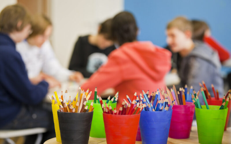Im Vordergrund sind bunte Becher mit Bleistiften und Kugelschreibern zu sehen, während im unscharfen Hintergrund eine Gruppe von Menschen zusammensitzt.