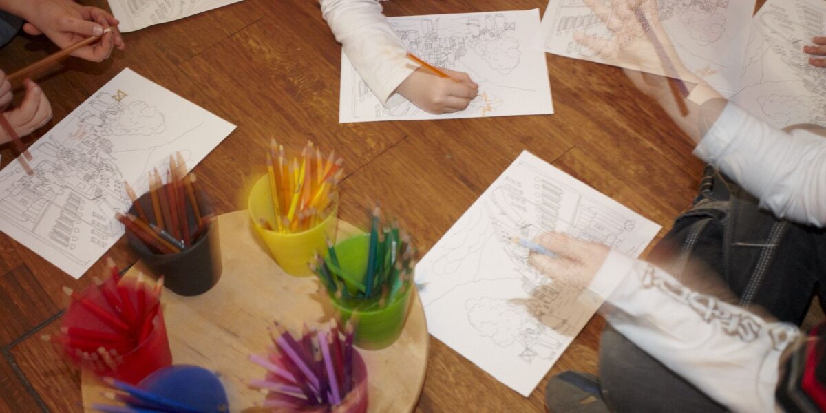 Die Kinder sitzen auf einem Holzboden und malen mit Buntstiften bedruckte Blätter aus, in der Mitte stehen Behälter mit Bleistiften auf einem Tablett.