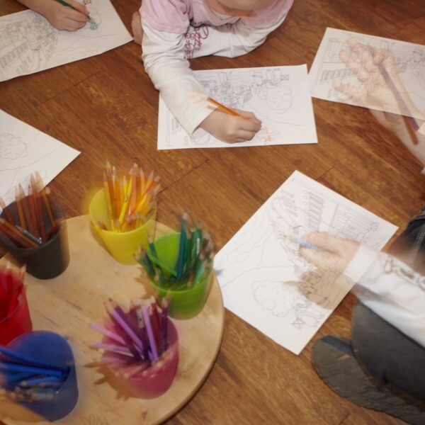 Die Kinder sitzen auf einem Holzboden und malen mit Buntstiften bedruckte Blätter aus, in der Mitte stehen Behälter mit Bleistiften auf einem Tablett.