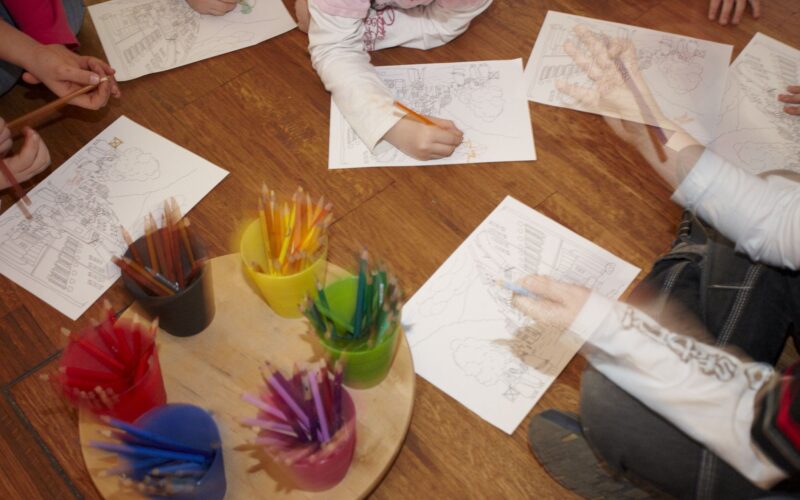 Die Kinder sitzen auf einem Holzboden und malen mit Buntstiften bedruckte Blätter aus, in der Mitte stehen Behälter mit Bleistiften auf einem Tablett.