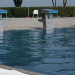 Drei nummerierte Startblöcke (1, 2, 3) sind am Rande eines Freibads zu sehen, mit klarem blauen Wasser und einer Hecke im Hintergrund.