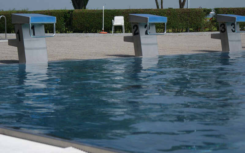 Drei nummerierte Startblöcke (1, 2, 3) sind am Rande eines Freibads zu sehen, mit klarem blauen Wasser und einer Hecke im Hintergrund.