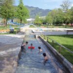 Mehrere Kinder spielen in einem flachen Wasserkanal in einem Park. Im Hintergrund sind Grasflächen, Bäume, Bänke und eine Wasserrutsche zu sehen.
