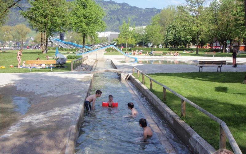 Mehrere Kinder spielen in einem flachen Wasserkanal in einem Park. Im Hintergrund sind Grasflächen, Bäume, Bänke und eine Wasserrutsche zu sehen.