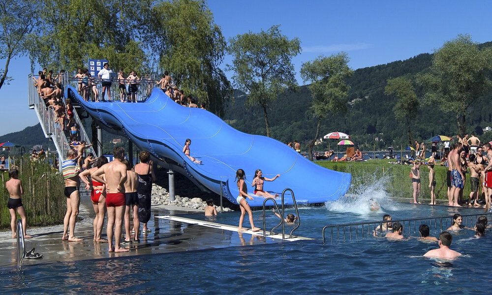 Menschen genießen einen sonnigen Tag in einem überfüllten Freibad mit einer großen blauen Dreifachrutsche, umgeben von Bäumen und Hügeln.