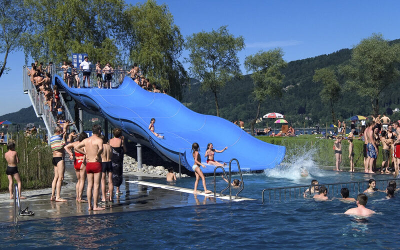 Menschen genießen einen sonnigen Tag in einem überfüllten Freibad mit einer großen blauen Dreifachrutsche, umgeben von Bäumen und Hügeln.