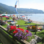 Menschen entspannen sich auf Liegestühlen und Handtüchern an einem grasbewachsenen Seeufer, mit bunten Sonnenschirmen, Blumen im Vordergrund und Bergen im Hintergrund.