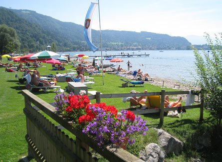 Menschen entspannen sich auf Liegestühlen und Handtüchern an einem grasbewachsenen Seeufer, mit bunten Sonnenschirmen, Blumen im Vordergrund und Bergen im Hintergrund.