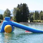 Menschen genießen eine blaue und gelbe aufblasbare Rutsche, die auf einem See schwimmt, umgeben von Bäumen und fernen Bergen.