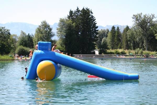 Menschen genießen eine blaue und gelbe aufblasbare Rutsche, die auf einem See schwimmt, umgeben von Bäumen und fernen Bergen.