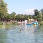 Eine Person schwimmt in einem See in der Nähe einer schwimmenden Rutsche und eines Spielgeräts, mit Gebäuden und Bäumen im Hintergrund unter einem klaren Himmel.