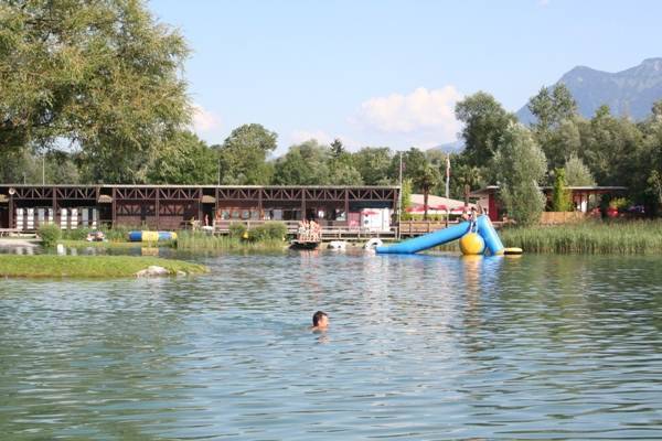 Eine Person schwimmt in einem See in der Nähe einer schwimmenden Rutsche und eines Spielgeräts, mit Gebäuden und Bäumen im Hintergrund unter einem klaren Himmel.