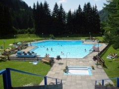 Ein von Gras und Bäumen umgebenes Freibad, in dem die Menschen schwimmen und sich auf Stühlen unter dem sonnigen Himmel entspannen.