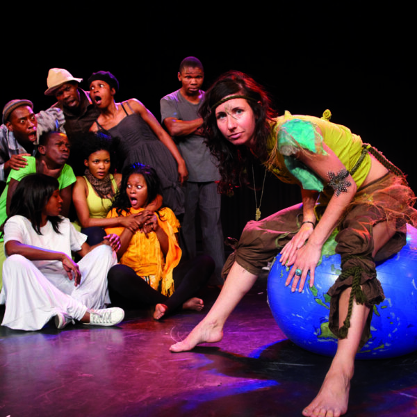 Eine Frau im Kostüm sitzt auf einem blauen Gymnastikball, der als Erde bemalt ist, während eine Gruppe von Menschen hinter ihr auf einer Bühne überrascht und besorgt reagiert.
