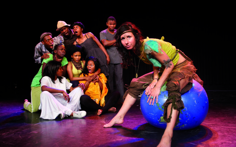 Eine Frau im Kostüm sitzt auf einem blauen Gymnastikball, der als Erde bemalt ist, während eine Gruppe von Menschen hinter ihr auf einer Bühne überrascht und besorgt reagiert.