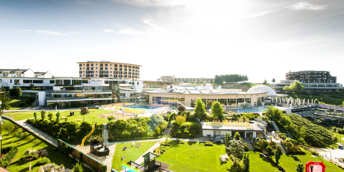 Ein großer Ferienkomplex mit mehreren Gebäuden, Außenpools, Gärten, Rasenflächen und Erholungsgebieten unter einem hellen, sonnigen Himmel.