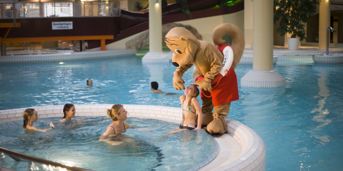 Eine Person in einem Eichhörnchen-Maskottchen-Kostüm steht am Rande eines Hallenbades und unterhält sich mit einem Kind, während andere in der Nähe schwimmen.