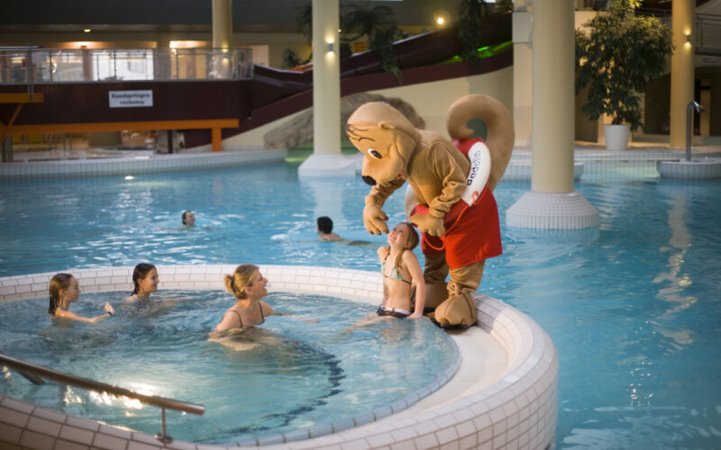 Eine Person in einem Eichhörnchen-Maskottchen-Kostüm steht am Rande eines Hallenbades und unterhält sich mit einem Kind, während andere in der Nähe schwimmen.