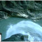 Ein Wasserlauf fließt schnell über Felsen in ein blaues Becken, das von rauem, felsigem Gelände umgeben ist.