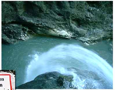 Ein Wasserlauf fließt schnell über Felsen in ein blaues Becken, das von rauem, felsigem Gelände umgeben ist.