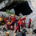 Eine Gruppe von Menschen in roten Anzügen und mit Helmen versammelt sich mit ihrer Ausrüstung am felsigen Eingang einer großen Höhle.