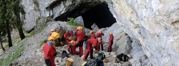Eine Gruppe von Menschen in roten Anzügen und mit Helmen versammelt sich mit ihrer Ausrüstung am felsigen Eingang einer großen Höhle.