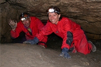 Zwei Personen in roten Overalls und mit Scheinwerfern kriechen durch einen engen Höhlengang und lächeln in die Kamera.
