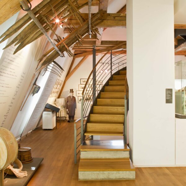 Innenraum eines modernen Museums mit Holzböden, sichtbaren Balken, Vitrinen, Wandausstellungen und einer Treppe, die in ein höheres Stockwerk führt.