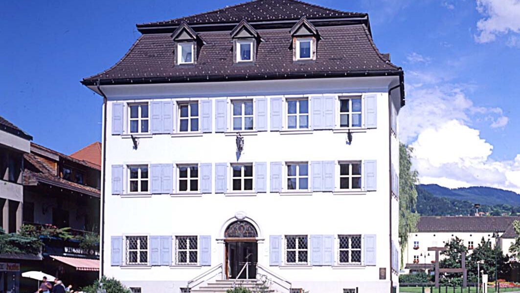 Ein weißes dreistöckiges Gebäude mit blauen Fensterläden, einem dunklen Ziegeldach und symmetrischen Fenstern, umgeben von Blumen und Grünpflanzen unter einem teilweise bewölkten Himmel.