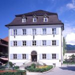 Ein weißes dreistöckiges Gebäude mit blauen Fensterläden, einem dunklen Ziegeldach und symmetrischen Fenstern, umgeben von Blumen und Grünpflanzen unter einem teilweise bewölkten Himmel.