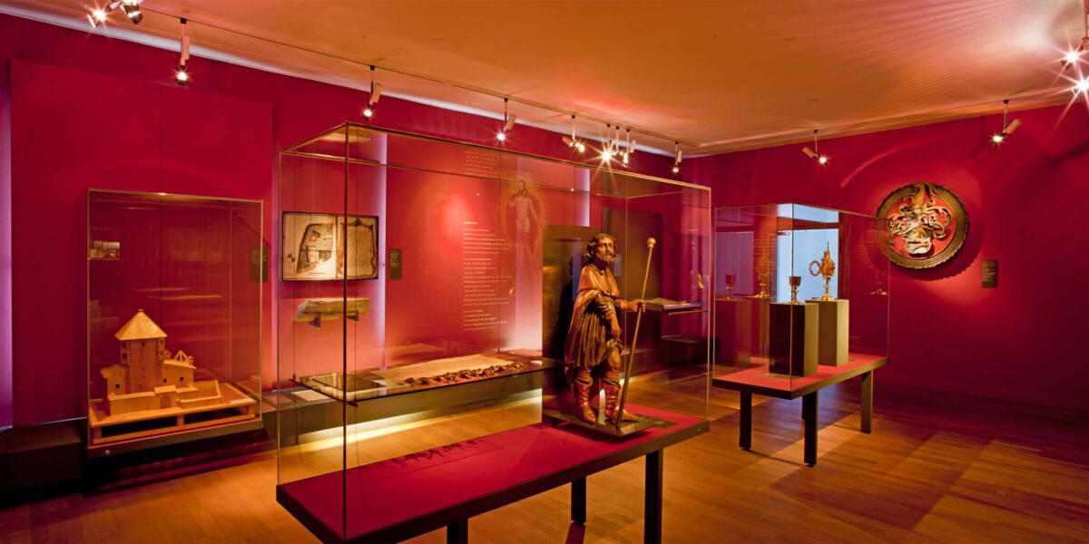 Eine Museumsgalerie mit roten Wänden zeigt Artefakte und Skulpturen in Glasvitrinen, die von Scheinwerfern beleuchtet werden.
