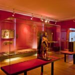 Eine Museumsgalerie mit roten Wänden zeigt Artefakte und Skulpturen in Glasvitrinen, die von Scheinwerfern beleuchtet werden.