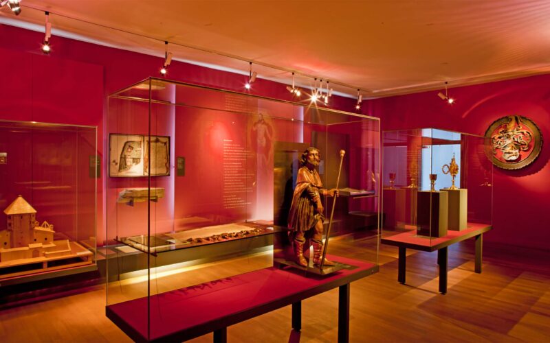 Eine Museumsgalerie mit roten Wänden zeigt Artefakte und Skulpturen in Glasvitrinen, die von Scheinwerfern beleuchtet werden.