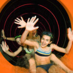 Drei Kinder in Badeanzügen verlassen eine orangefarbene Wasserrutsche, lächeln und winken aufgeregt mit den Händen.