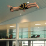 Eine Person in geblümter Badehose befindet sich in der Luft und taucht waagerecht über einem Hallenbad mit Balkonen und Liegestühlen im Hintergrund.