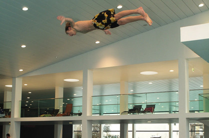Eine Person in geblümter Badehose befindet sich in der Luft und taucht waagerecht über einem Hallenbad mit Balkonen und Liegestühlen im Hintergrund.