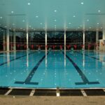 Ein Hallenbad mit Bahnmarkierungen und roten Fahnen, die über dem Wasser hängen; große Fenster säumen die Wände und die Deckenbeleuchtung spiegelt sich auf der Wasseroberfläche.