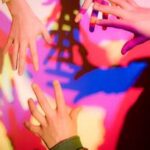 Vier Hände, die sich einander entgegenstrecken, mit bunten, sich überlappenden Schatten, die auf eine Wand im Hintergrund geworfen werden.