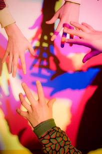 Vier Hände, die sich einander entgegenstrecken, mit bunten, sich überlappenden Schatten, die auf eine Wand im Hintergrund geworfen werden.