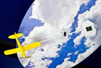 Ein gelbes Modellflugzeug hängt vor einer Himmelskulisse mit Wolken, die mit einem Stab an einem weißen wolkenförmigen Objekt befestigt ist.