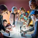Eine Gruppe von Kindern und Erwachsenen interagiert in einer wissenschaftlichen Ausstellung mit Licht und Schatten, indem sie Objekte auf einem runden beleuchteten Tisch benutzt.