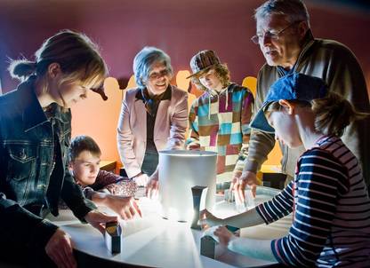 Eine Gruppe von Kindern und Erwachsenen interagiert in einer wissenschaftlichen Ausstellung mit Licht und Schatten, indem sie Objekte auf einem runden beleuchteten Tisch benutzt.