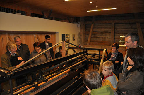 Eine Gruppe von Personen beobachtet die Vorführung einer historischen Maschine in einem Holzgebäude, deren Funktion von einem Ausbilder erklärt wird.