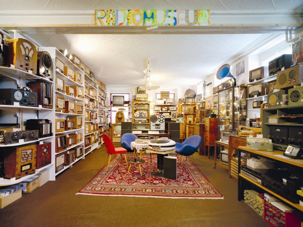 Ein Raum voller alter Radios und Audiogeräte in Regalen, mit einem runden Tisch und bunten Stühlen auf einem roten Teppich in der Mitte; darüber ein Schild mit der Aufschrift "RADIO MUSEUM".