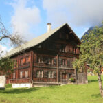 Ein großes Holzhaus mit mehreren Fenstern steht auf einem grasbewachsenen Rasen unter einem teilweise bewölkten Himmel, in dessen Nähe sich ein paar Bäume befinden.